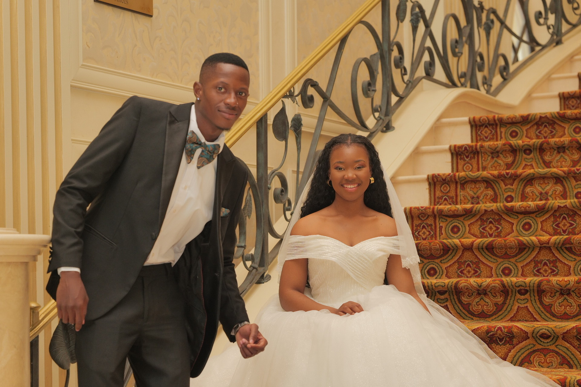 A smiling bride and groom pose on the carpeted grand staircase at The Grand Palm, Gaborone